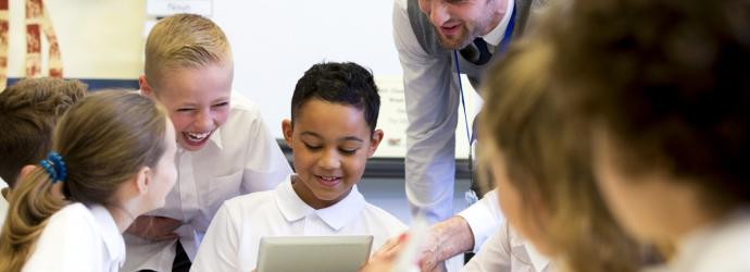 a male teacher sits supervising a group of children who are working on whiteboards and digita