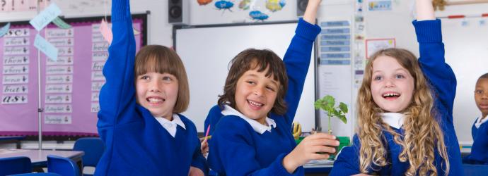 Group of 3 primary school age children enthusiastically putting their hand up sat around a classroom desk, there are small plant trays, felt tip pens and paper on the table. 