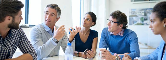 Group of professionals sat at a table having a discussion, there is water and note pads on the table with one person taking down notes. 