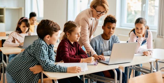 A teacher and four students all looking at a laptop in a classroom setting