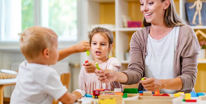 An older woman with two young children. She is teaching them to play with blocks, and they are in a library setting