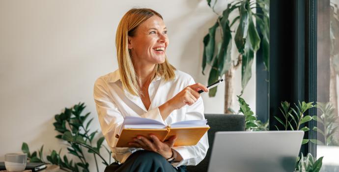 A smiling young professional is sitting in front of a laptop. She has an open book in her hand and is surrounded by plants