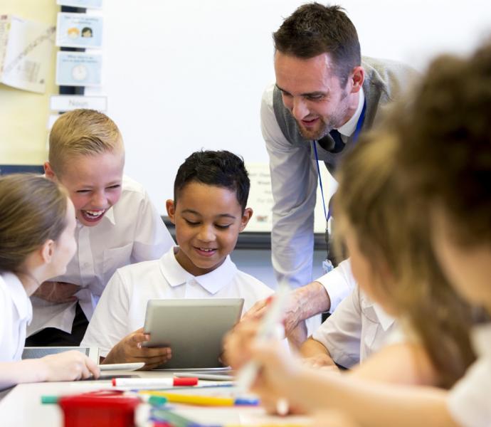 a male teacher sits supervising a group of children who are working on whiteboards and digita