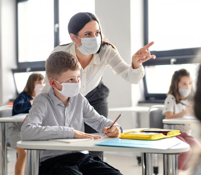 Female teacher supporting male pupil  - both are wearing masks in the classroom