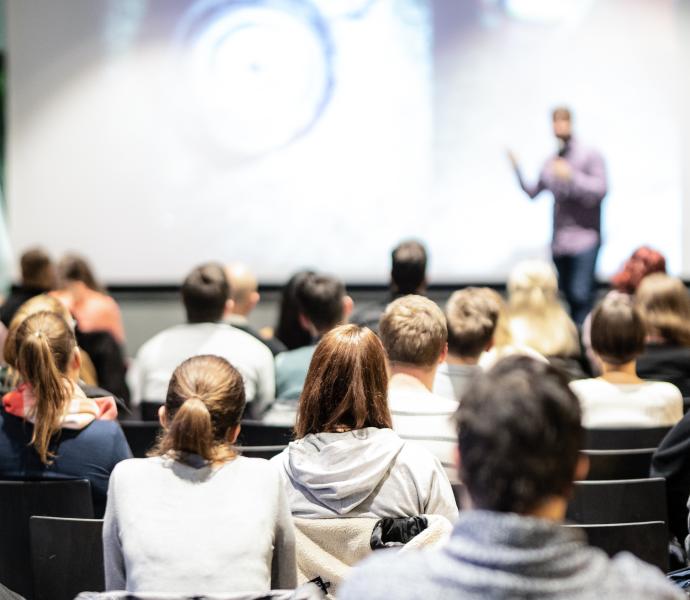 Group of professionals sat in a conference setting with a speaker standing at the front delivering content 