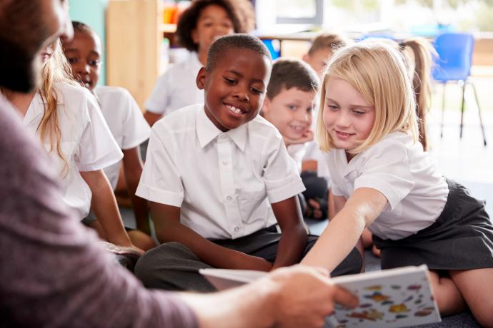 Primary school children sat on the carpet in group reading with a teacher holding out a book