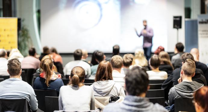 Group of professionals sat in a conference setting with a speaker standing at the front delivering content 